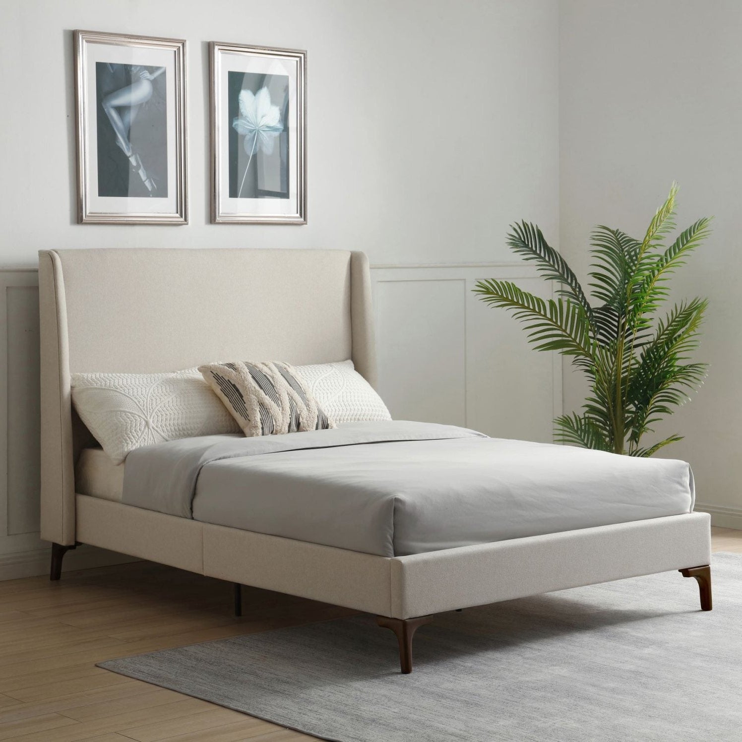 A modern upholstered bed frame in a bright, minimalist bedroom. The bed features a tall, wingback-style headboard and matching low-profile footboard in a light beige fabric, supported by dark wood legs.