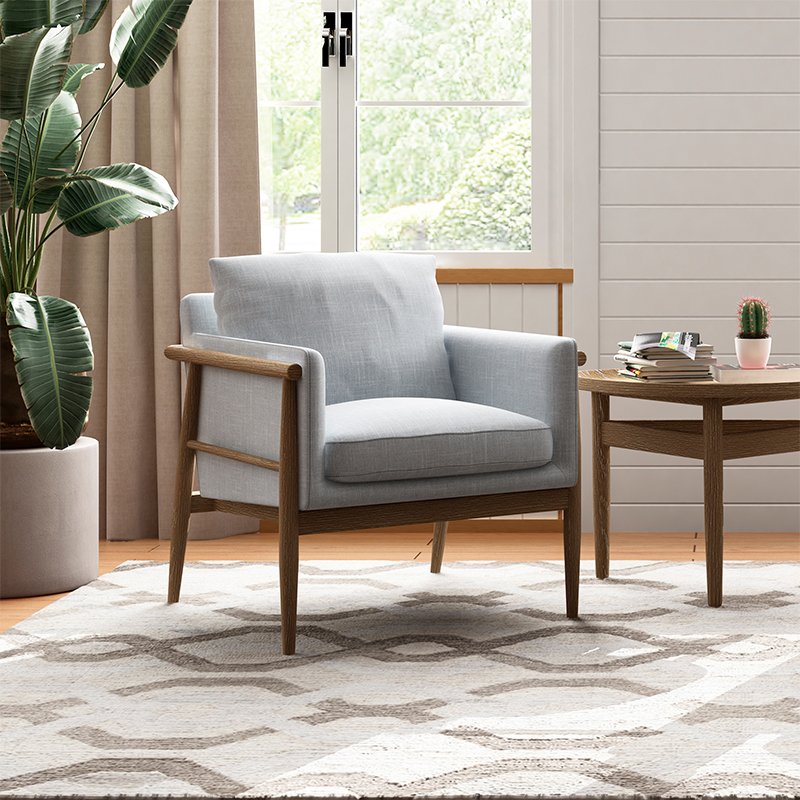 A grey / blue upholstered armchair with a wooden frame sits in a bright living room setting next to a matching wooden side table, with a large potted plant and a patterned rug in the background.