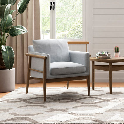 A grey / blue upholstered armchair with a wooden frame sits in a bright living room setting next to a matching wooden side table, with a large potted plant and a patterned rug in the background.