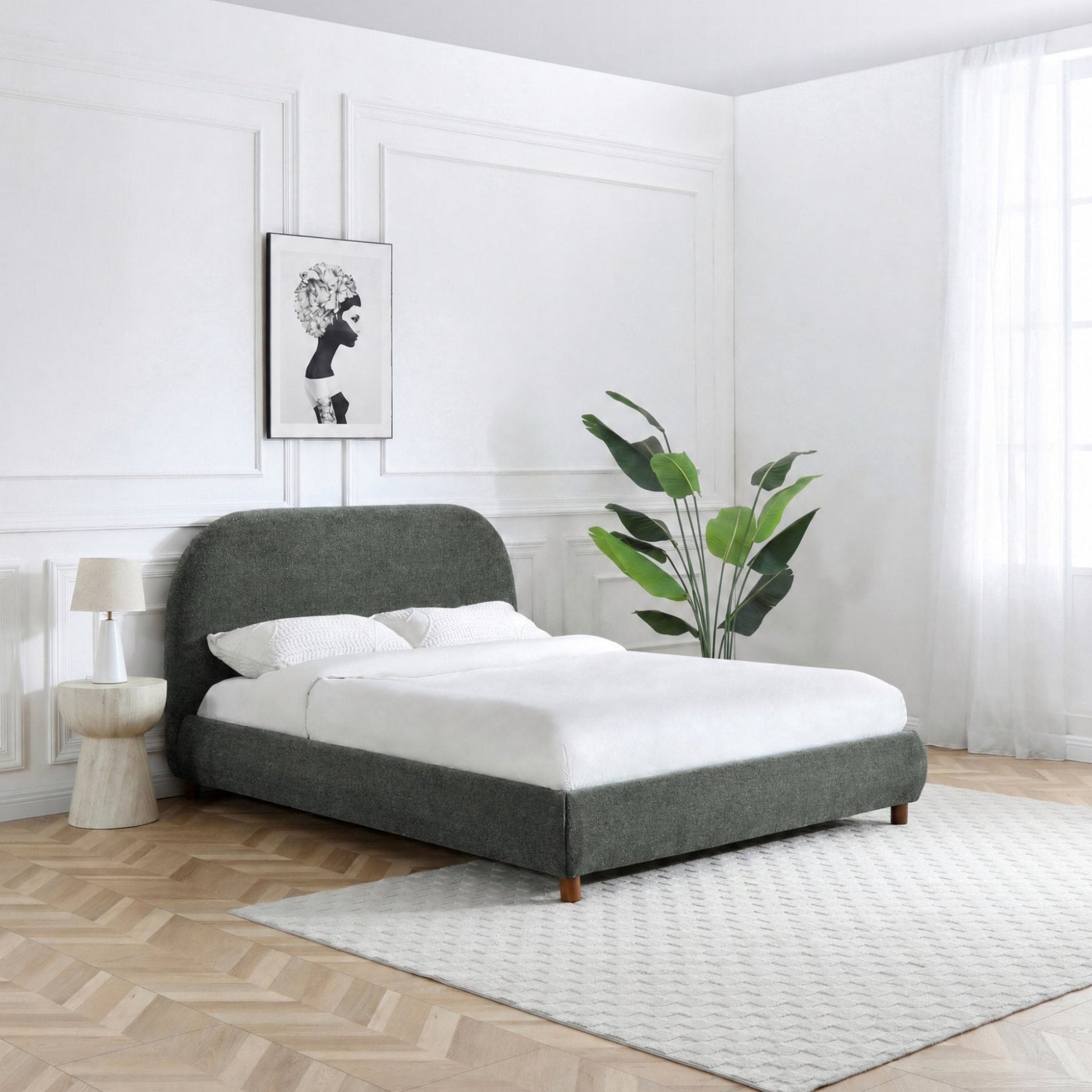 A modern upholstered bed frame in dark gray fabric, styled with white bedding in a bright, minimalist bedroom with white paneled walls, a light wood floor, and a white patterned rug.