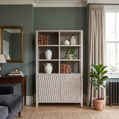 A tall white wooden bookcase with open upper shelves and textured paneled lower doors, styled in a living room with books, white ceramic vases, a small potted plant, beige curtains, a window, and a potted fiddle-leaf fig.