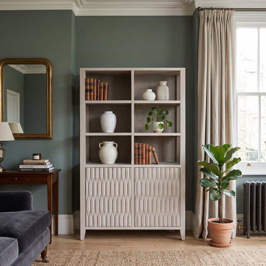 A tall white wooden bookcase with open upper shelves and textured paneled lower doors, styled in a living room with books, white ceramic vases, a small potted plant, beige curtains, a window, and a potted fiddle-leaf fig.