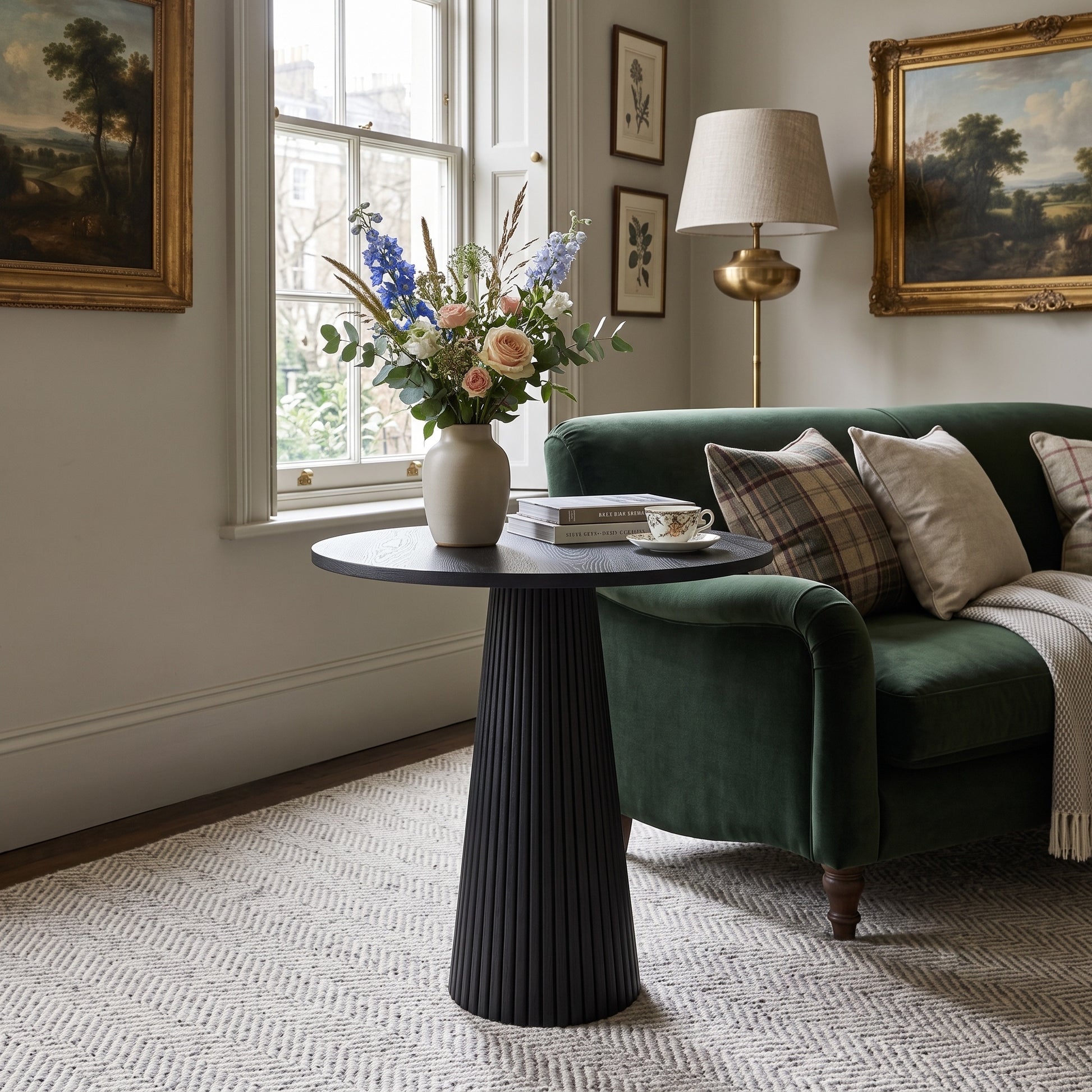 A dark round side table with a fluted pedestal base in a styled living room, decorated with a vase of flowers, books, and a teacup. The table is positioned next to a green velvet sofa with plaid and cream pillows.