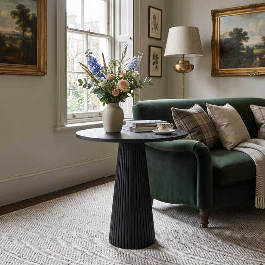 A dark round side table with a fluted pedestal base in a styled living room, decorated with a vase of flowers, books, and a teacup. The table is positioned next to a green velvet sofa with plaid and cream pillows.