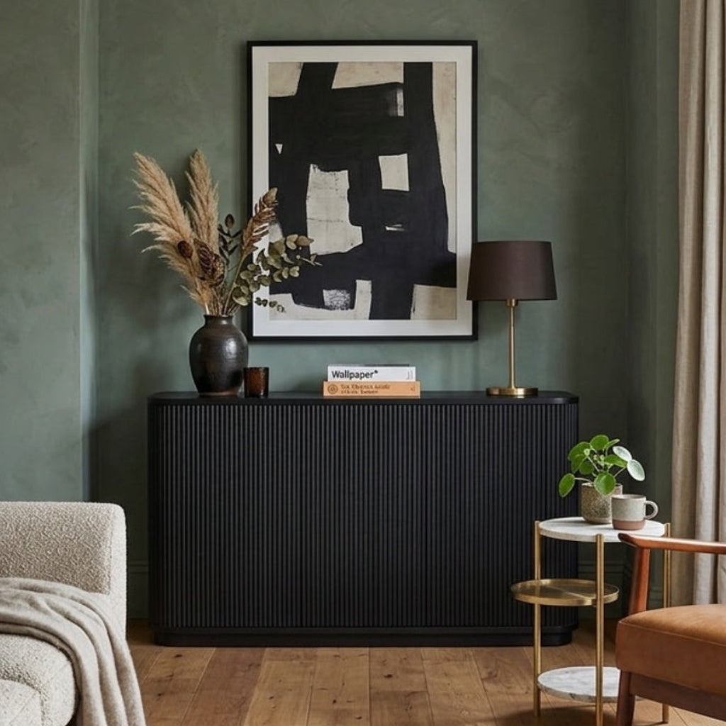 A styled living room interior featuring a dark charcoal ribbed sideboard console against a textured sage green wall, with a black-framed abstract print above. The room includes a beige textured sofa, a mid-century modern brown leather armchair, a small gold side table with a plant, and a window with beige curtains.
