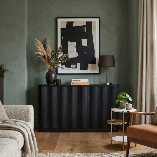 A styled living room interior featuring a dark charcoal ribbed sideboard console against a textured sage green wall, with a black-framed abstract print above. The room includes a beige textured sofa, a mid-century modern brown leather armchair, a small gold side table with a plant, and a window with beige curtains.