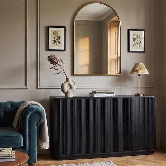 A styled interior room featuring a tall black pleated sideboard or credenza against a beige paneled wall, beneath a gold-framed arched mirror. A teal velvet sofa, round wood coffee table, textured rug, and warm natural light from a window are visible in the room.