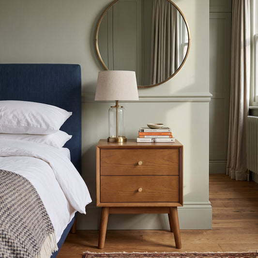 A mid-century modern wooden nightstand with two drawers and gold knobs, styled in a bedroom next to a bed with a blue headboard, a table lamp, and a round gold-framed mirror on a sage green wall.