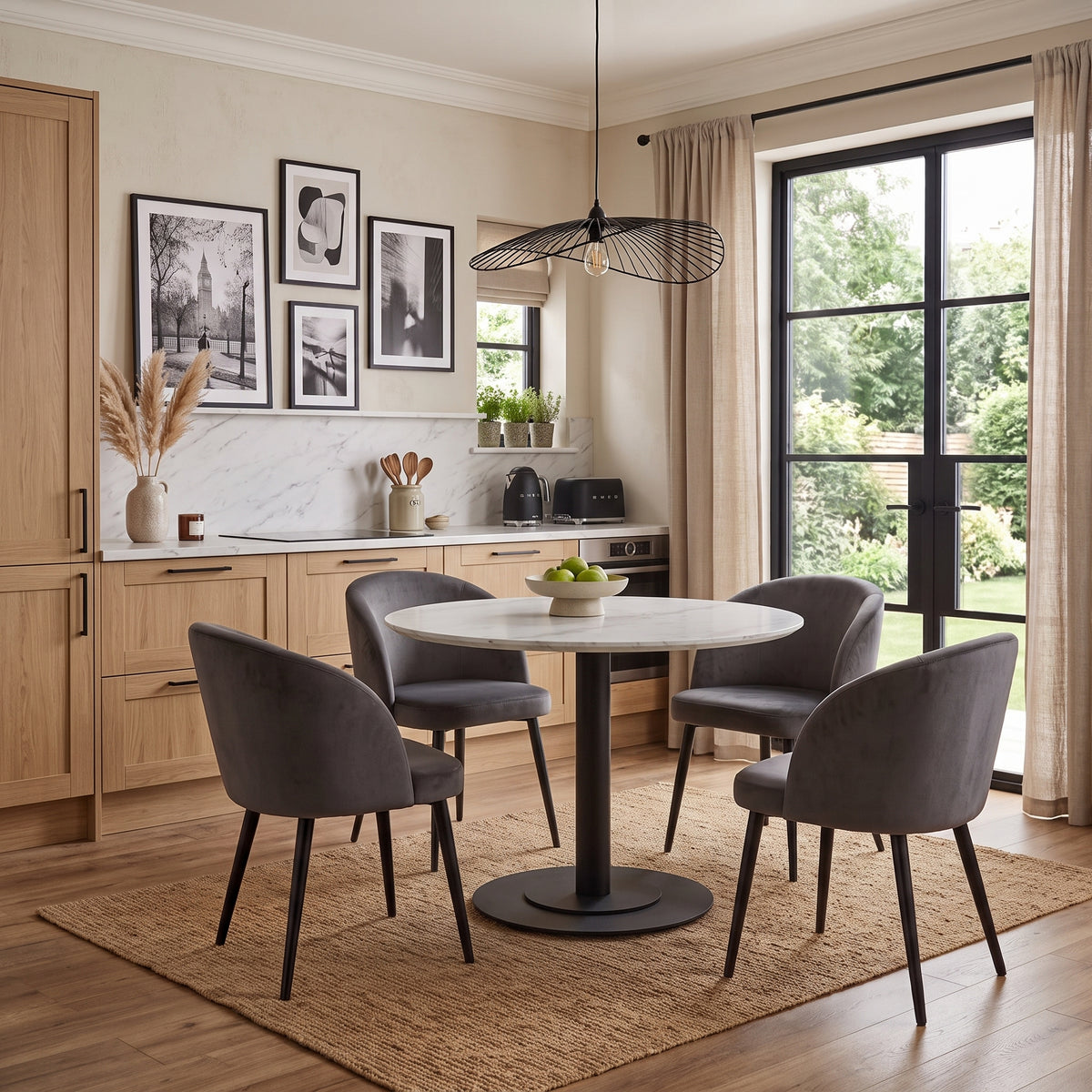 A modern dining set featuring a round white marble-top table with a black pedestal base, surrounded by four gray velvet upholstered chairs with black legs, set on a natural jute rug in a bright, contemporary kitchen-dining room.