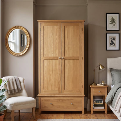 A light oak double-door wardrobe with a single drawer beneath, shown in a styled bedroom setting next to a bed and nightstand.