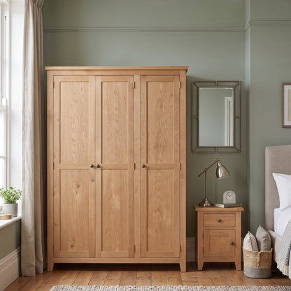 A light natural oak three-door wardrobe in a styled bedroom with sage green walls, a wooden nightstand, a bed, and a textured rug.