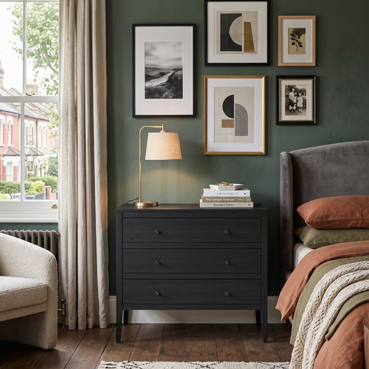 A styled bedroom interior featuring a dark charcoal gray three-drawer nightstand centered against a deep green wall, decorated with framed art and a table lamp. The room includes a bed with rust-colored bedding, a gray upholstered headboard, a window with beige curtains, and a light-colored armchair.