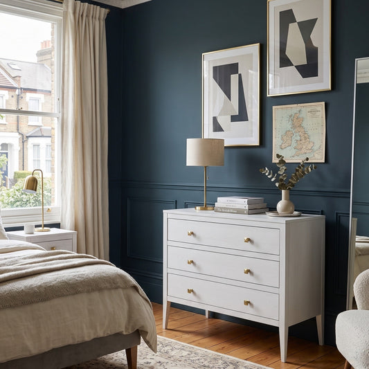 A styled bedroom interior with a dark blue wall, a white three-drawer dresser, framed abstract wall art, a bed, window, curtains, and a mirror.