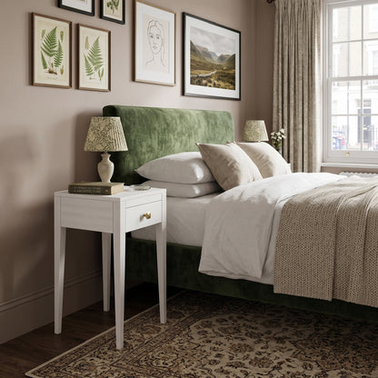 A styled bedroom scene featuring a bed with a prominent green velvet headboard, white bedding, a white nightstand with a patterned table lamp, and framed botanical and landscape prints on a beige wall.