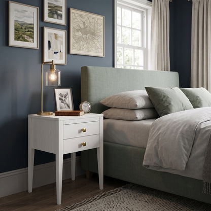 A styled bedroom scene featuring a white two-drawer nightstand with gold hardware next to a light sage green upholstered bed. The room has dark navy blue walls, framed art, a brass table lamp, and a window with curtains.