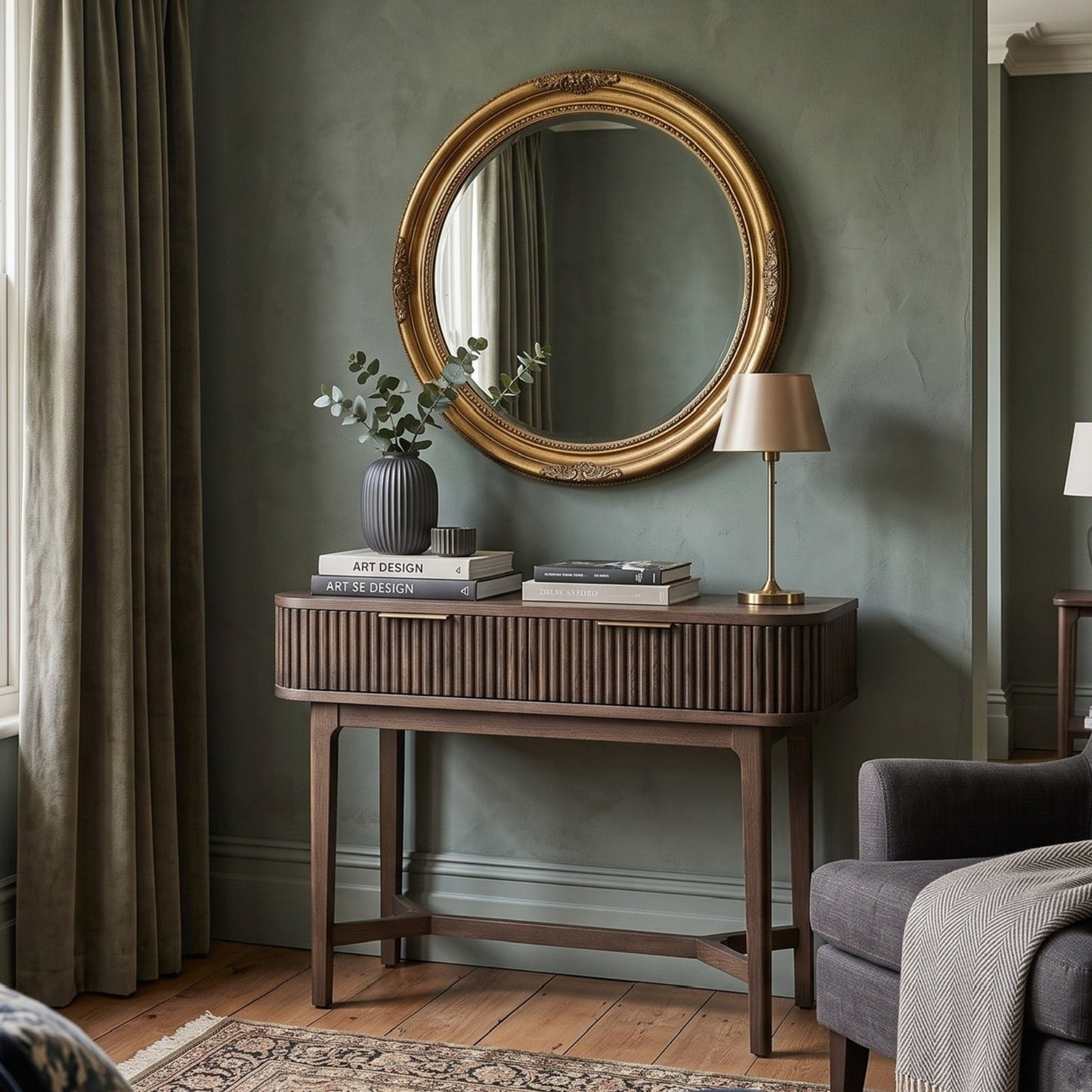 A styled interior room scene featuring a round ornate gold-framed mirror mounted on a textured sage green wall above a dark wood console table. 
