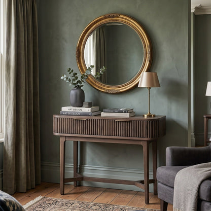 A styled interior room scene featuring a round ornate gold-framed mirror mounted on a textured sage green wall above a dark wood console table. 