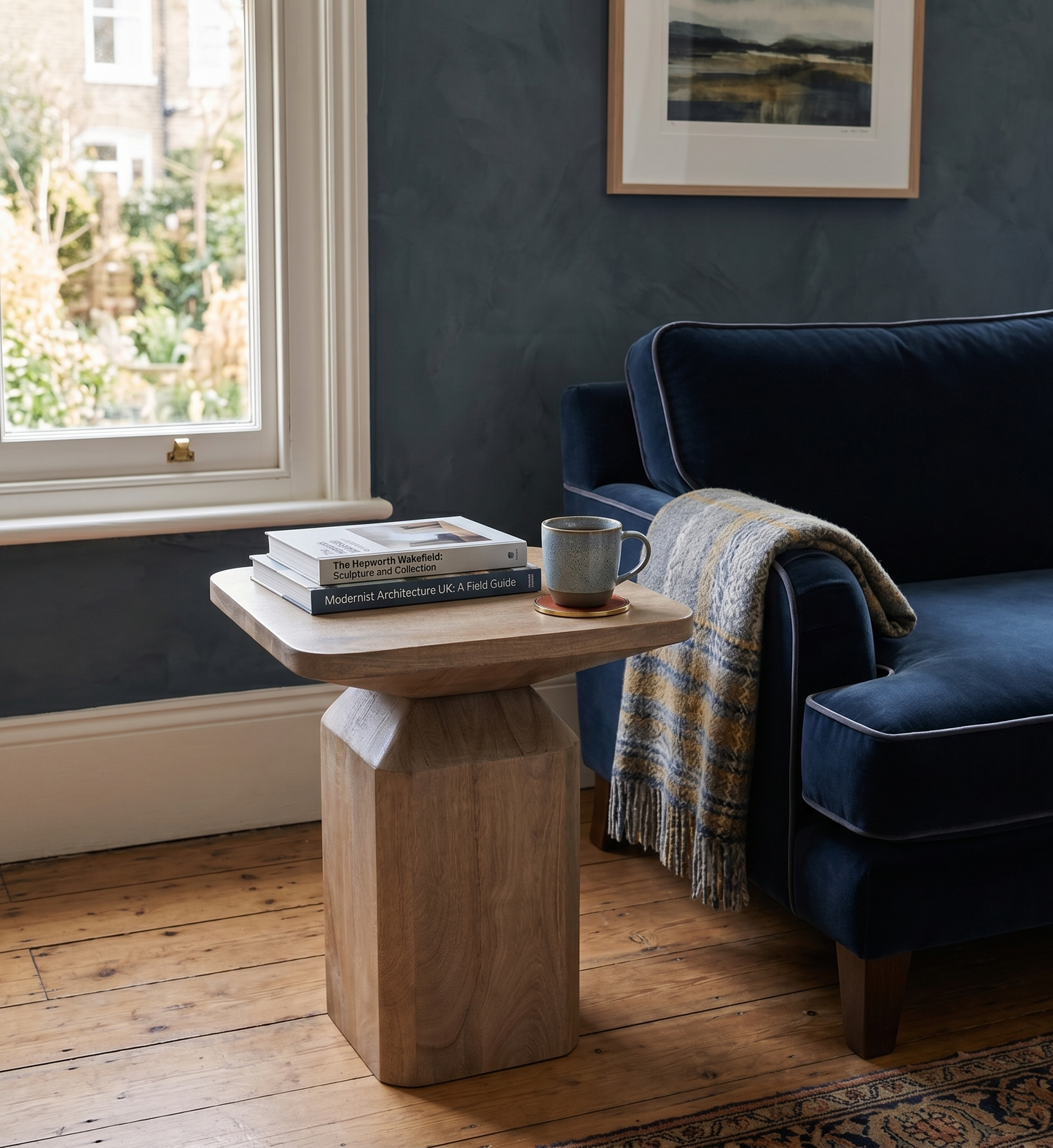 Navy blue sofa with a wooden side table and books in a room with a window and framed picture on the wall.