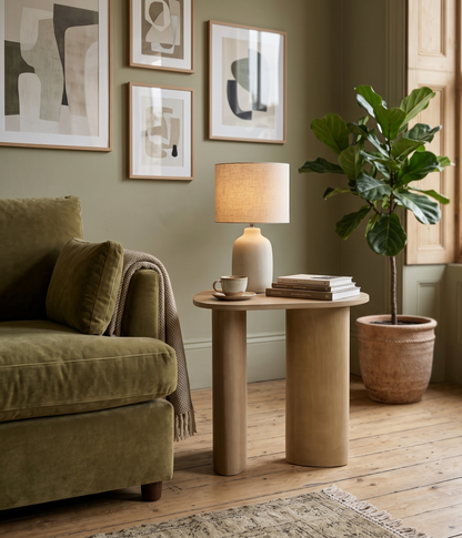 A round wooden side table in a styled living room setting, styled with a beige ceramic table lamp, a stack of books, and a small white teacup and saucer. The table is positioned next to a green velvet armchair and a potted fiddle-leaf fig plant.