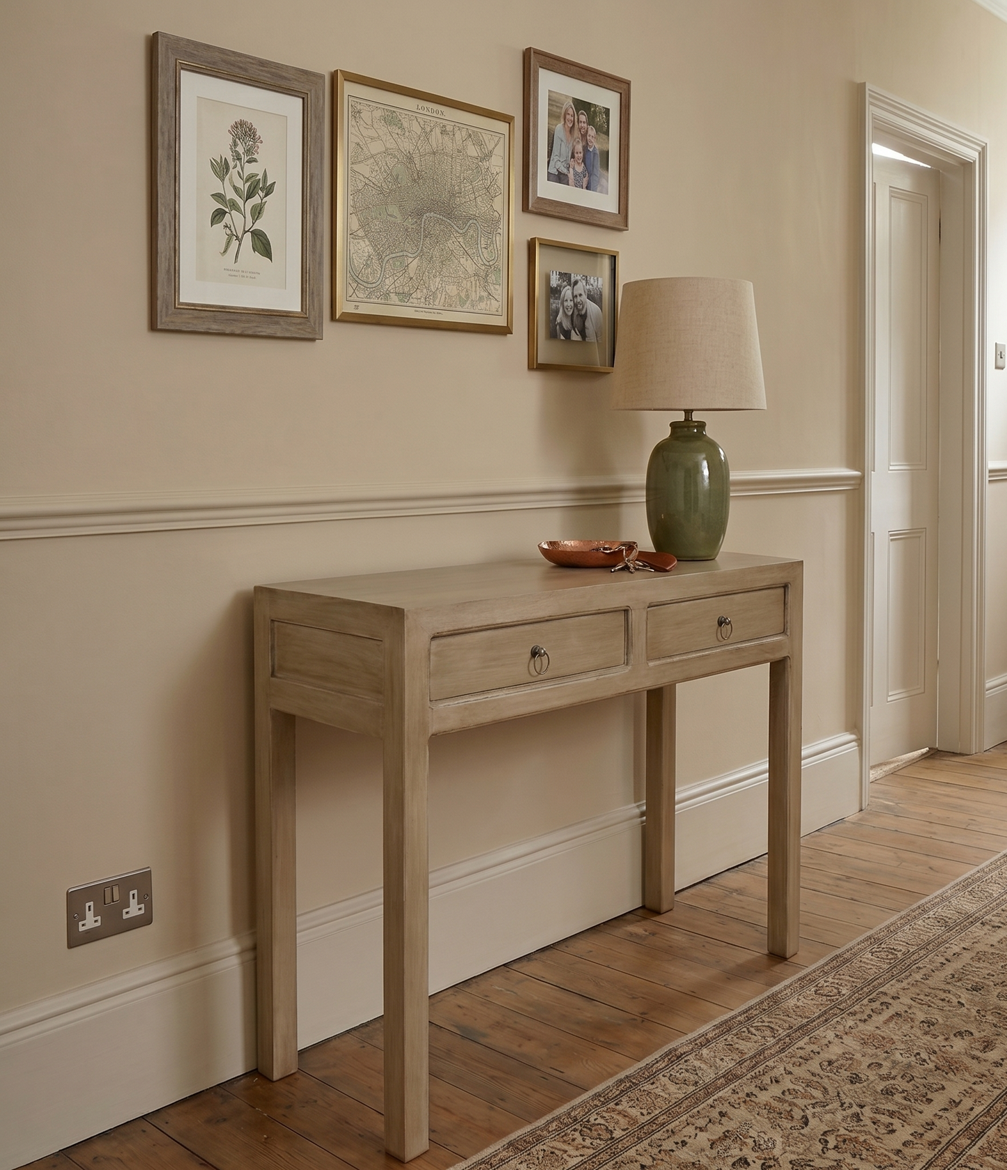 A light natural wood console table with two drawers, styled in a hallway with a green ceramic lamp, framed prints, and a patterned rug.