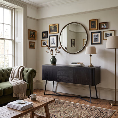 A styled living room interior featuring a dark wood sideboard console with a large round mirror, a green velvet armchair with a cream throw, a wooden coffee table, and framed artwork on a light wall.