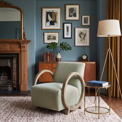 A styled interior room scene featuring a light sage green armchair with a circular wooden frame, set on a textured rug next to a brass side table and a floor lamp. The room has blue-green walls, a wooden fireplace, and framed botanical art.