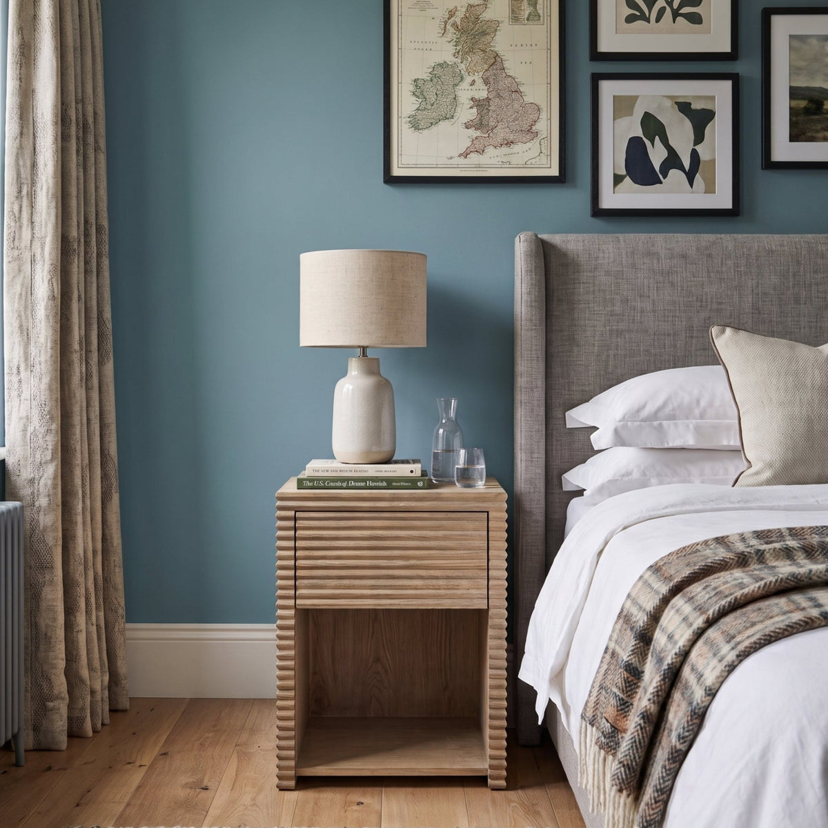 A styled bedroom scene featuring a light oak ribbed nightstand with a white ceramic table lamp, set against a slate blue wall with framed art and next to a bed with a gray upholstered headboard.