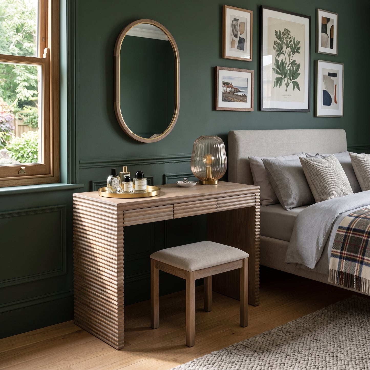 A wooden vanity table with a slatted front and matching stool in a styled bedroom setting next to a bed with green walls and framed artwork.