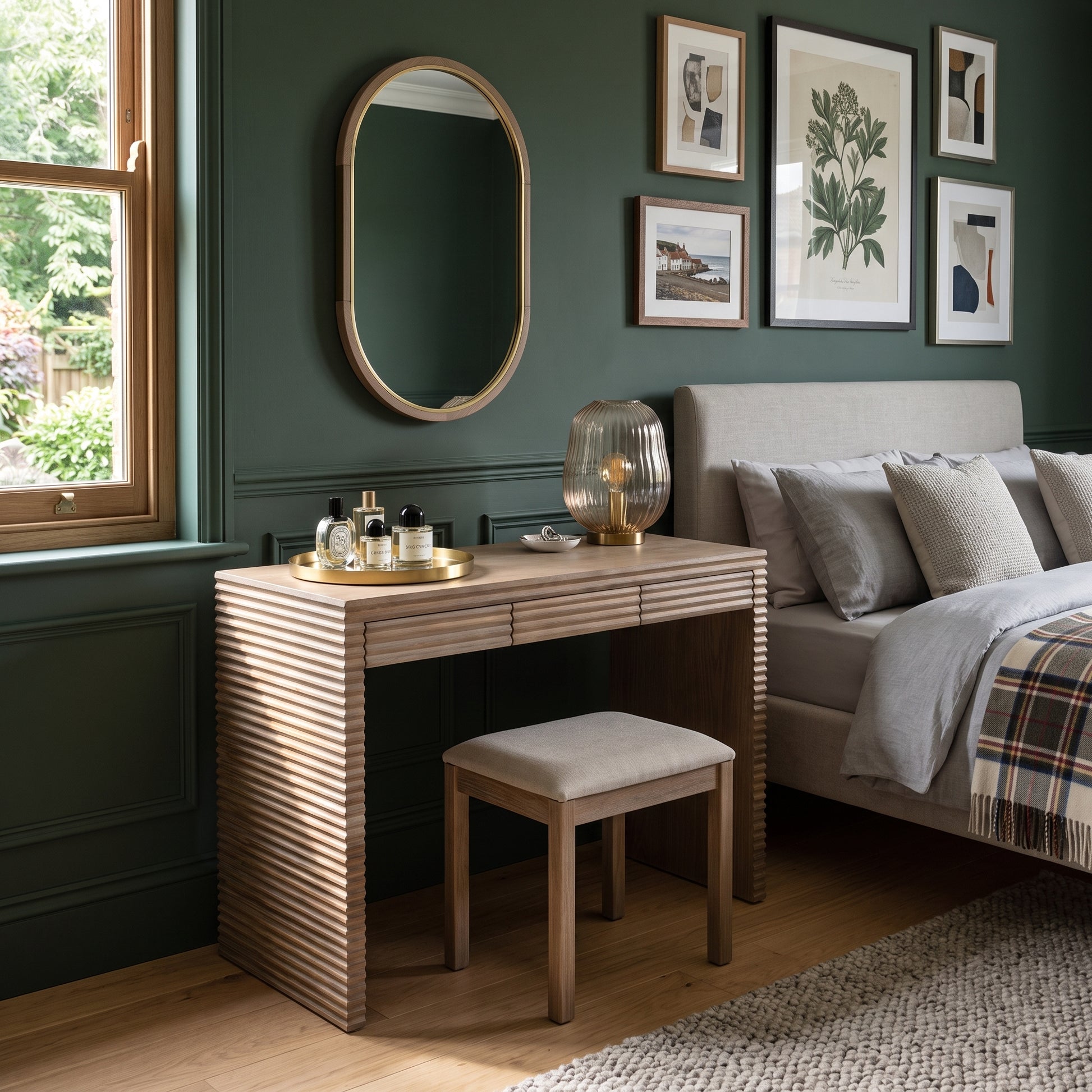 A wooden vanity table with a slatted front and matching stool in a styled bedroom setting next to a bed with green walls and framed artwork.