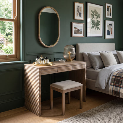 A wooden vanity table with a slatted front and matching stool in a styled bedroom setting next to a bed with green walls and framed artwork.