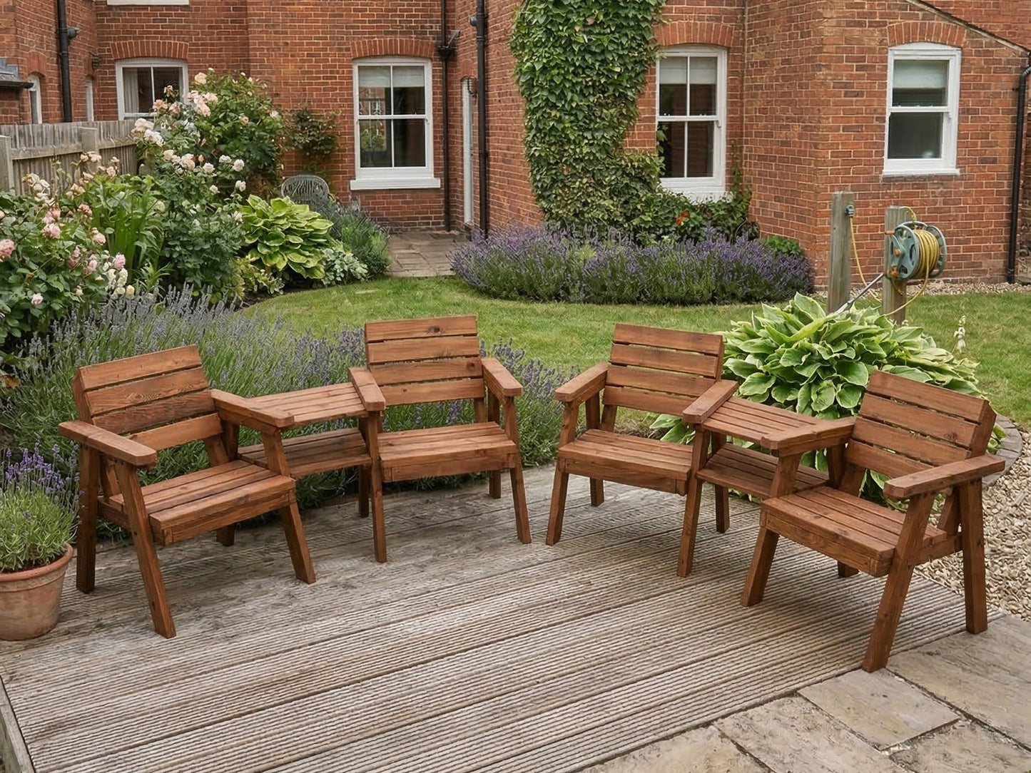 Wooden garden furniture set on a wooden deck in front of a brick building with garden plants.